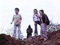 Three children stand on a mound of earth looking shocked, with a sinister, hooded figure behind them.